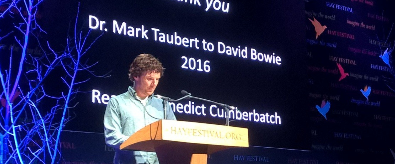 A man is reading from a lectern on a stage, The screen behind him says: "Thank you, Mark Taubert to David Bowie 2016, Benedict Cumberbatch